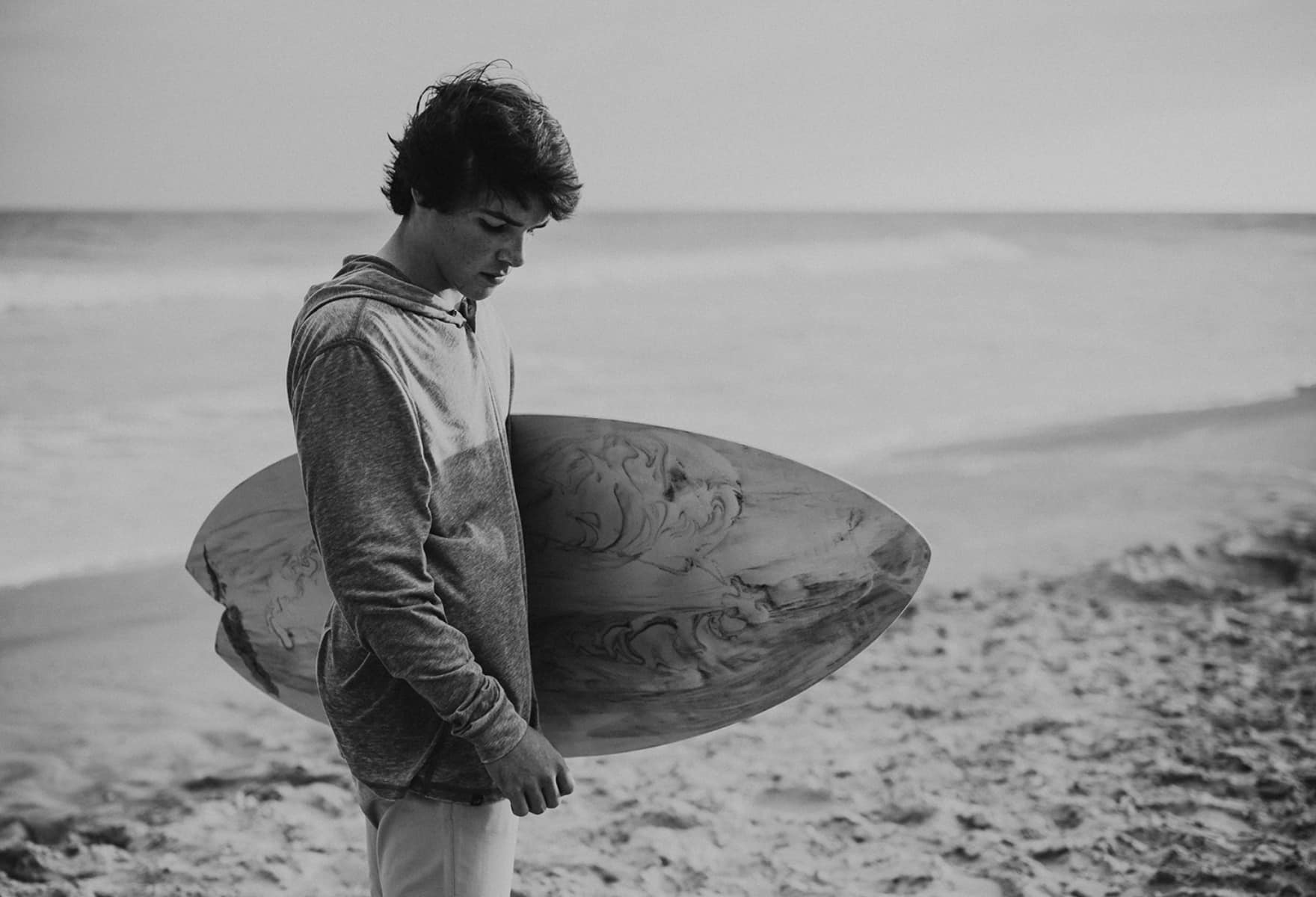 Young surfer holding a surfboard on the beach, gazing down at the sand, with the ocean in the background